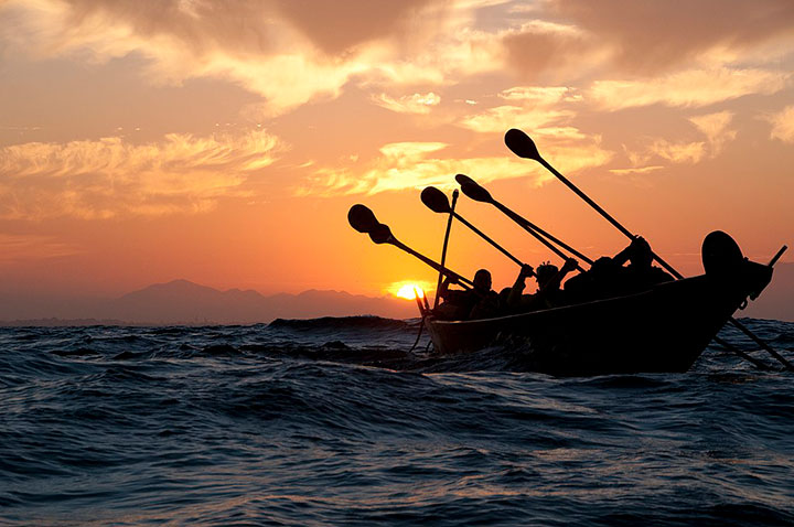 people paddling a long boat on the ocean at sunset